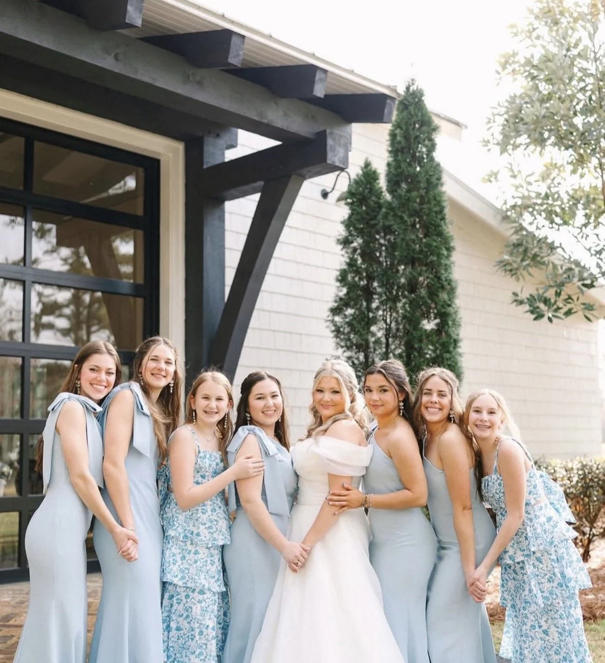 A group of women, including a bride and bridesmaids, standing outdoors in front of a modern house with trees in the background, dressed in elegant light blue and patterned dresses, smiling and holding hands.
