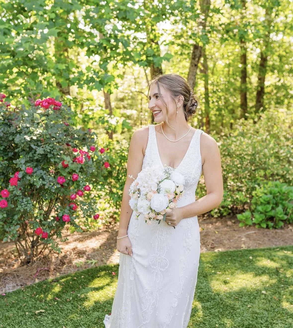 A bride stands outdoors in a garden, wearing a white wedding dress and holding a bouquet of white and pink flowers, smiling and looking to her left.