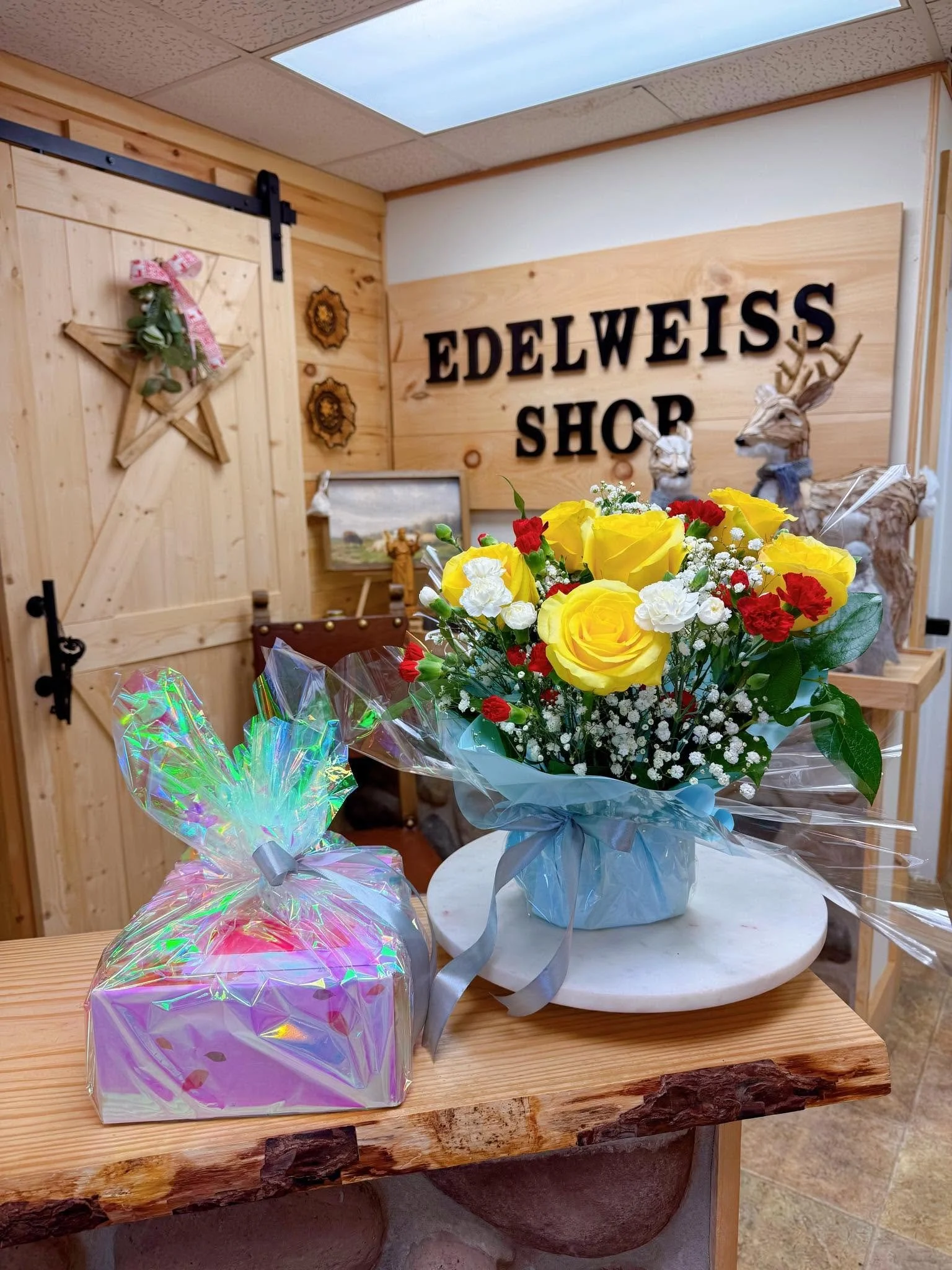 A bouquet of yellow, red, and white flowers wrapped in light blue paper on a marble stand in front of a wooden wall with the sign 'Edelweiss Shop' and decorated wooden animals and star with greenery.