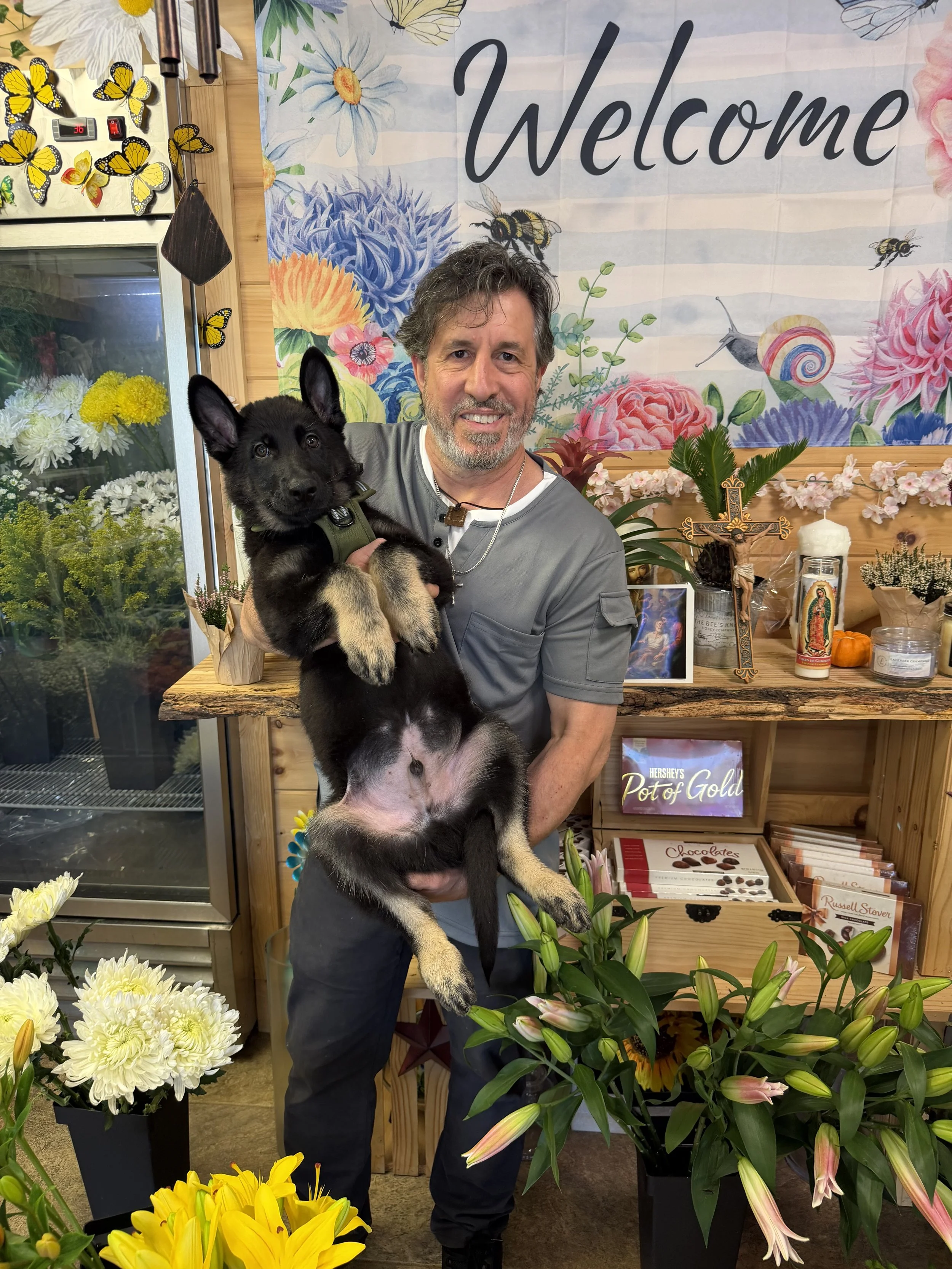 A man with gray hair and beard holding a black and tan puppy in a flower shop. The background has a colorful floral and bee-themed banner with the word 'Welcome'. There are various flowers in pots in the foreground, including yellow, white, pink, and purple blooms. The shop has a wooden counter with religious and decorative items, chocolates, and books.