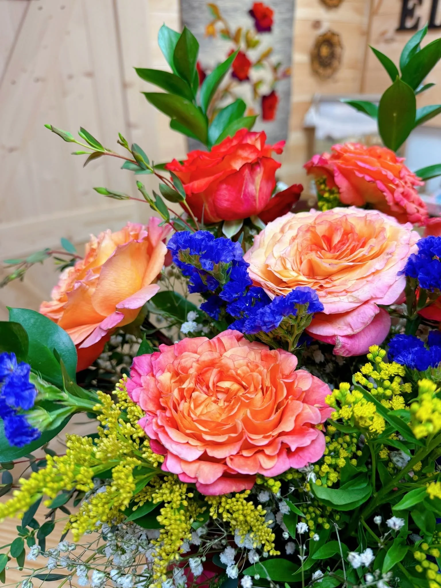 A vibrant bouquet of mixed flowers including orange roses, pink carnations, blue statice, yellow solidago, and small white blossoms, with green leaves and stems, set indoors with a wooden wall background.