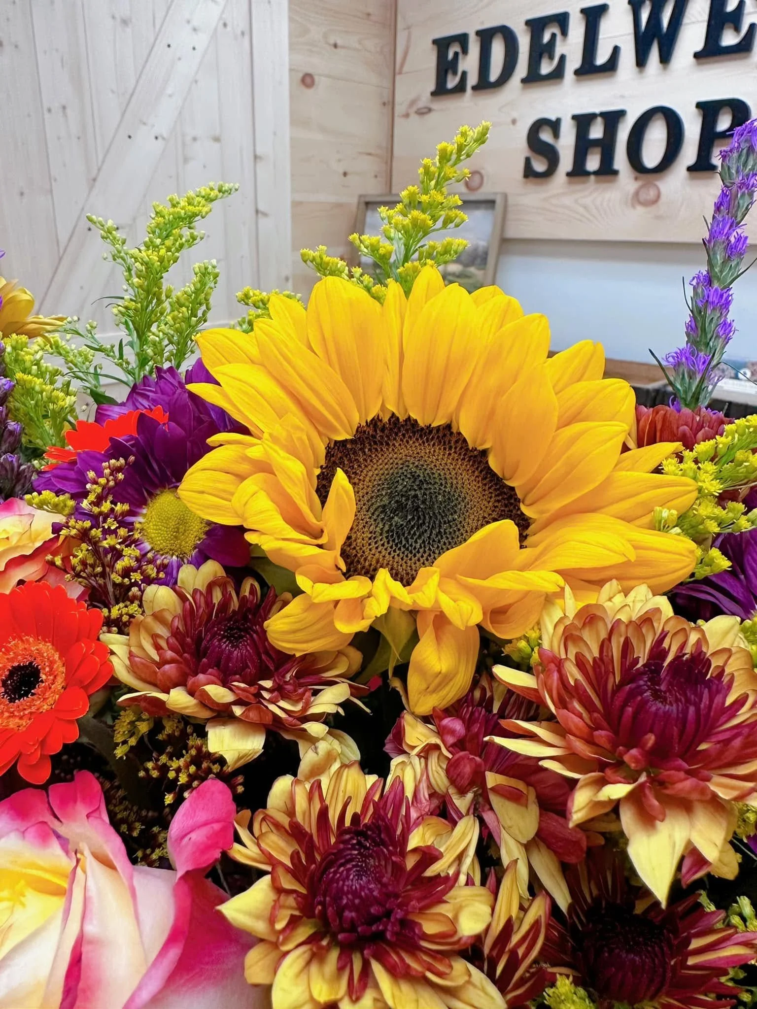 Close-up of a bouquet of colorful flowers including a large yellow sunflower at the center, surrounded by purple, red, pink, and orange flowers, with a light wooden wall and a sign that reads 'EDELWEIS' in the background.
