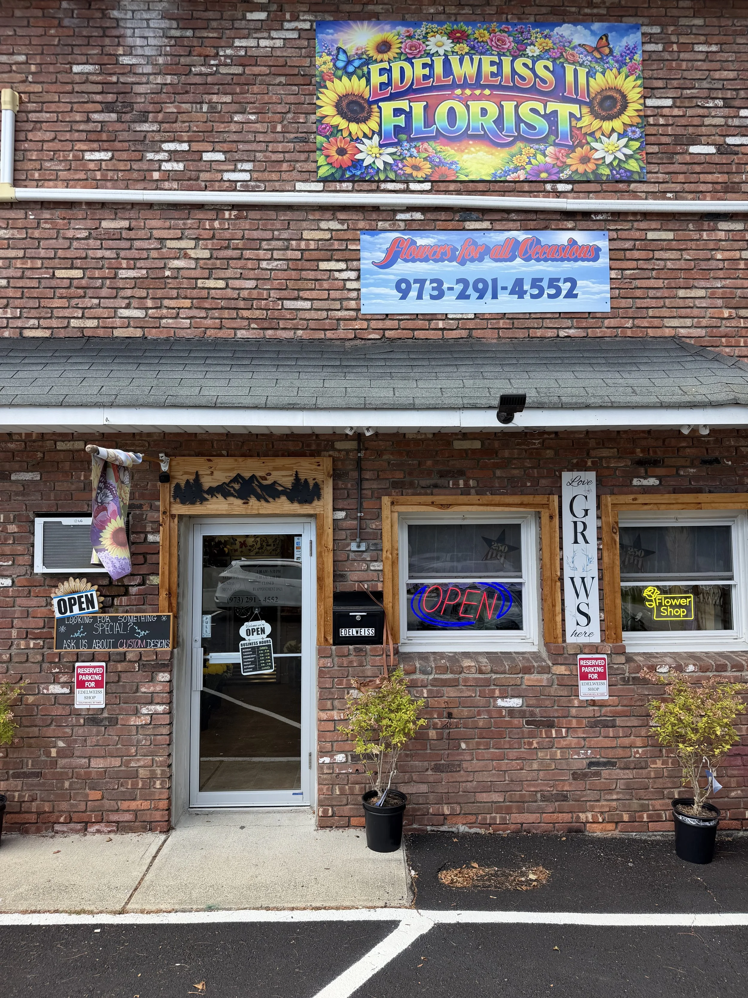 The image shows the storefront of Edelweiss II Florist. The building has a brick exterior with decorative wooden trim around the windows. There are several signs, including a colorful sign at the top with sunflower and butterfly graphics that reads 'EDELWEISS II FLORIST,' and a smaller blue sign below that says 'flowers for all Occasions' with a phone number. The door has an 'OPEN' neon sign, and there are plants in pots outside. The sidewalk and parking lot are in front of the shop.