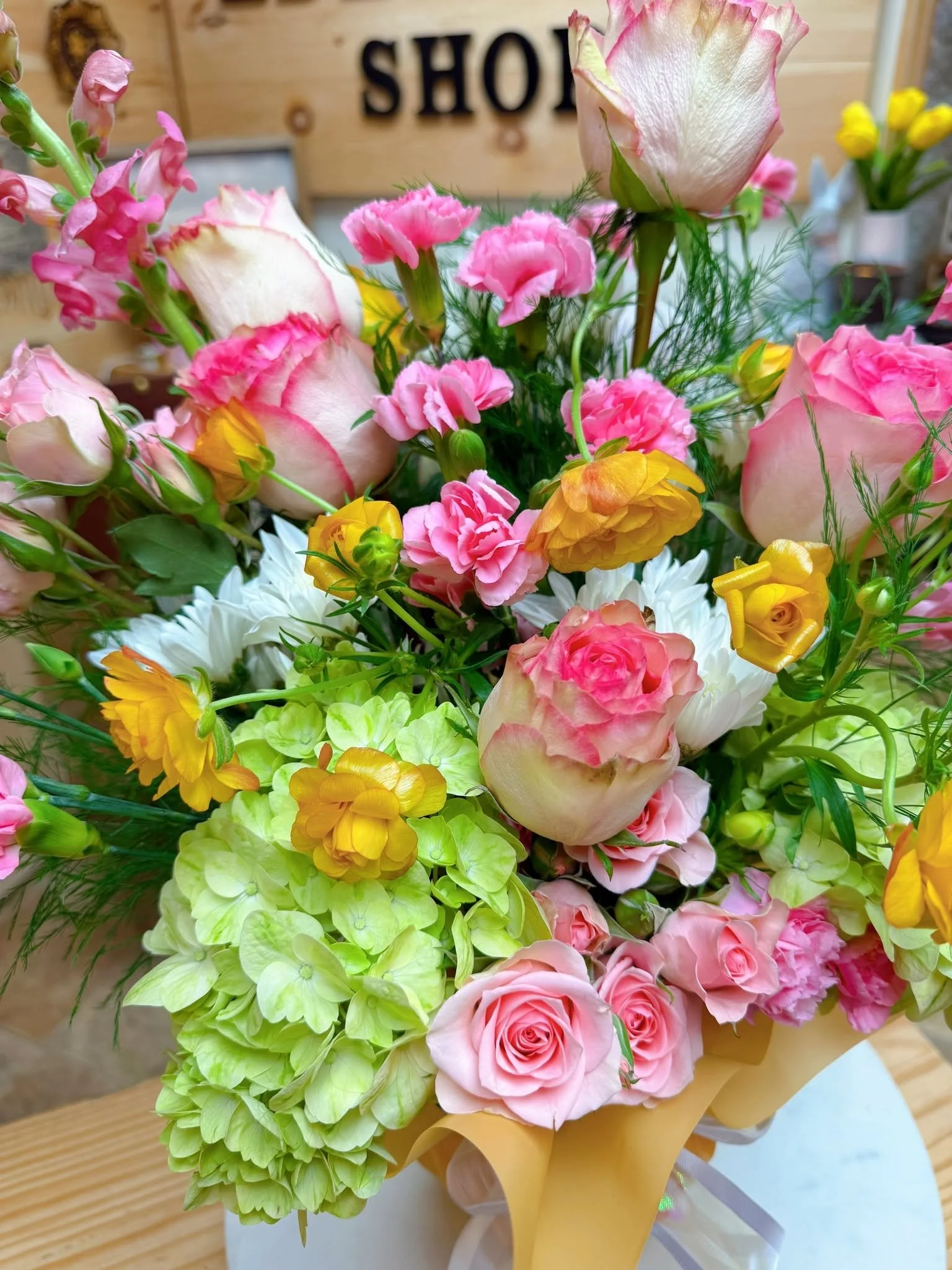 A colorful bouquet of pink roses, yellow and orange flowers, white hydrangeas, and greenery on a wooden table.