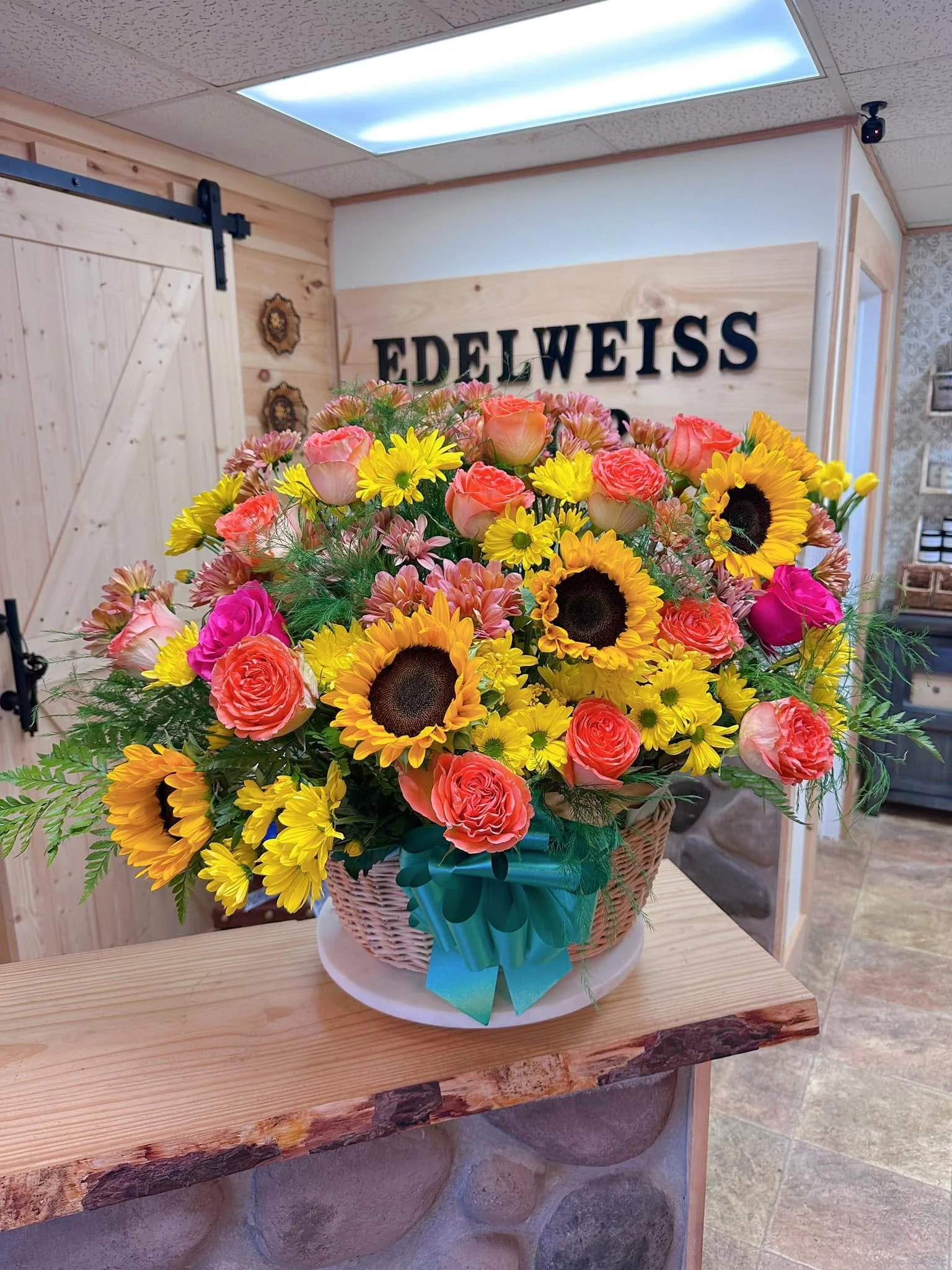 A colorful flower bouquet featuring pink roses, yellow sunflowers, and assorted smaller flowers in a woven basket with a teal ribbon inside a shop with a wooden sign that says 'Edelweiss'.