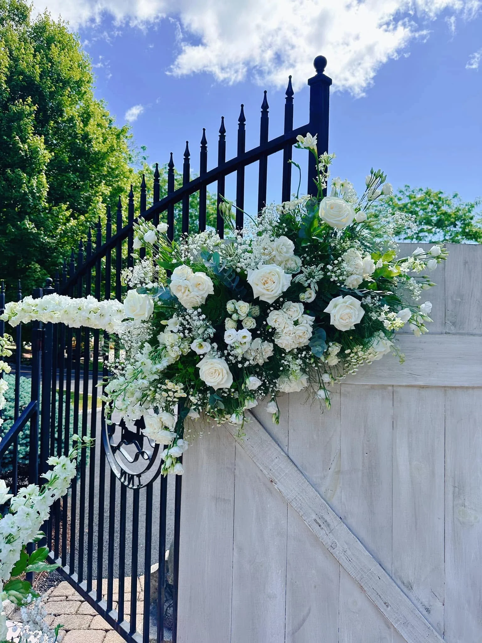 White floral arrangement with roses and baby's breath attached to a black wrought-iron fence and stone wall, with a blue sky and green trees in the background.