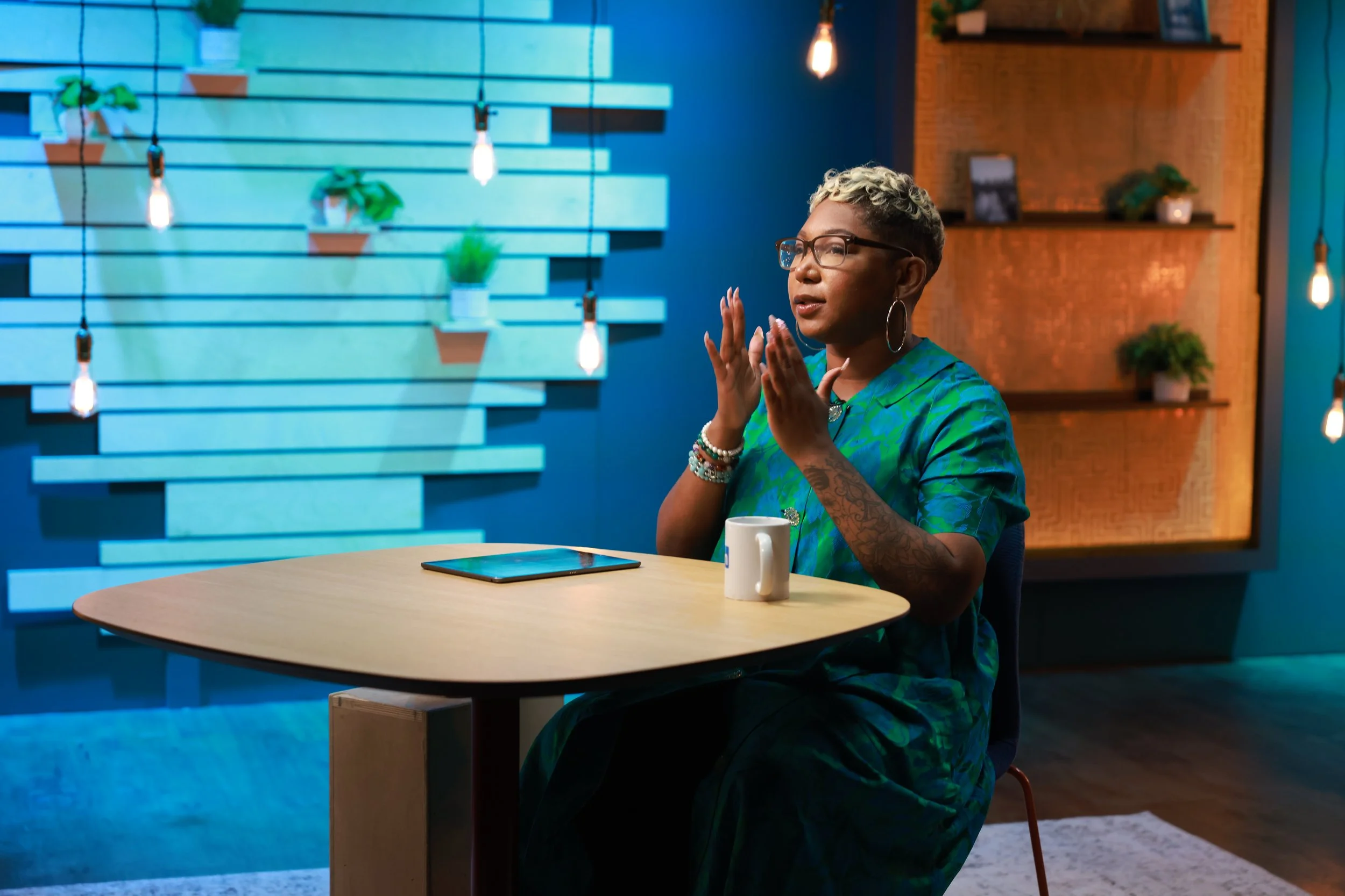 A woman with short curly blonde hair, glasses, and multiple bracelets, wearing a green patterned top, sits at a round wooden table with a mug and tablet, in a modern, well-lit room with blue walls, hanging Edison bulbs, and decorative shelves with plants.