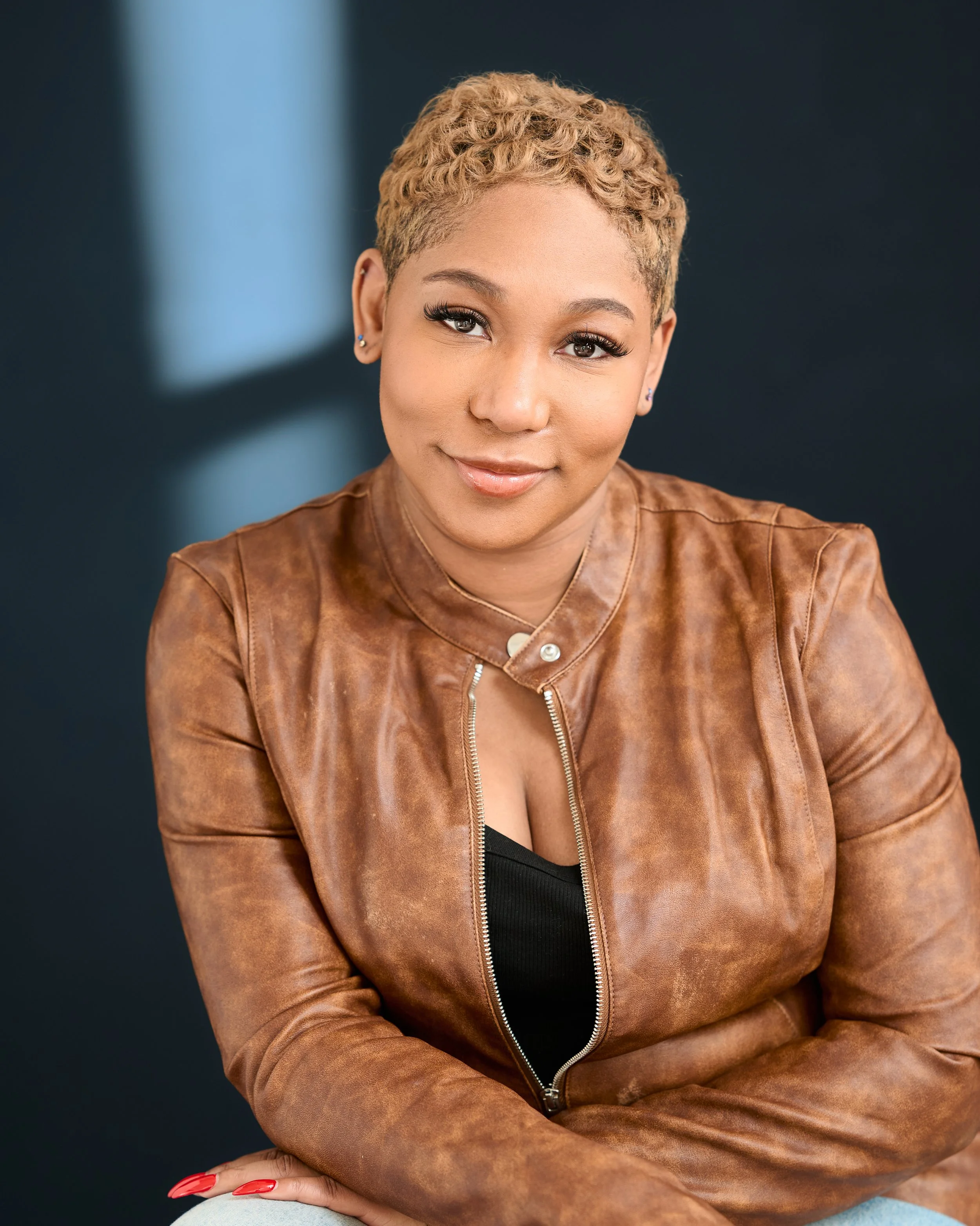 Jamé  with short, curly blonde hair and earrings smiling and wearing a brown leather jacket and black top against a dark background.