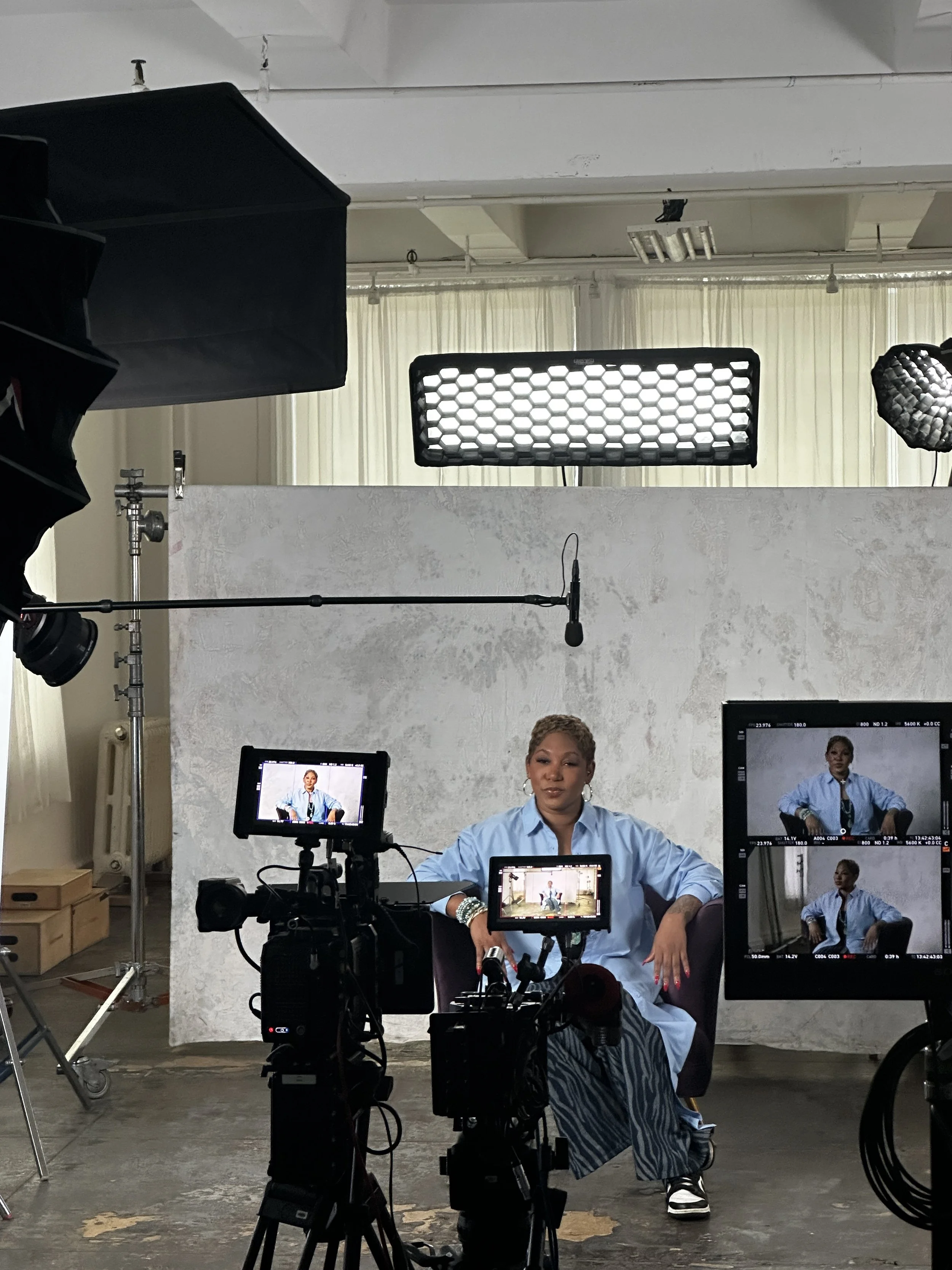 A woman sitting in front of a camera setup in a photography or filming studio, with multiple monitors and lights around her, appears to be hosting or participating in a video shoot.