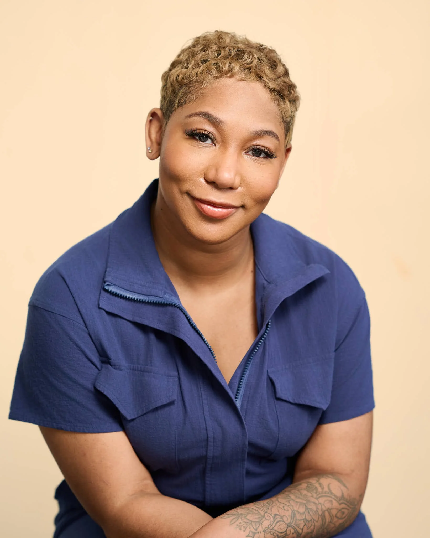 A woman with short curly blonde hair, wearing a blue zip-up shirt, is smiling gently with arms crossed, against a beige background.