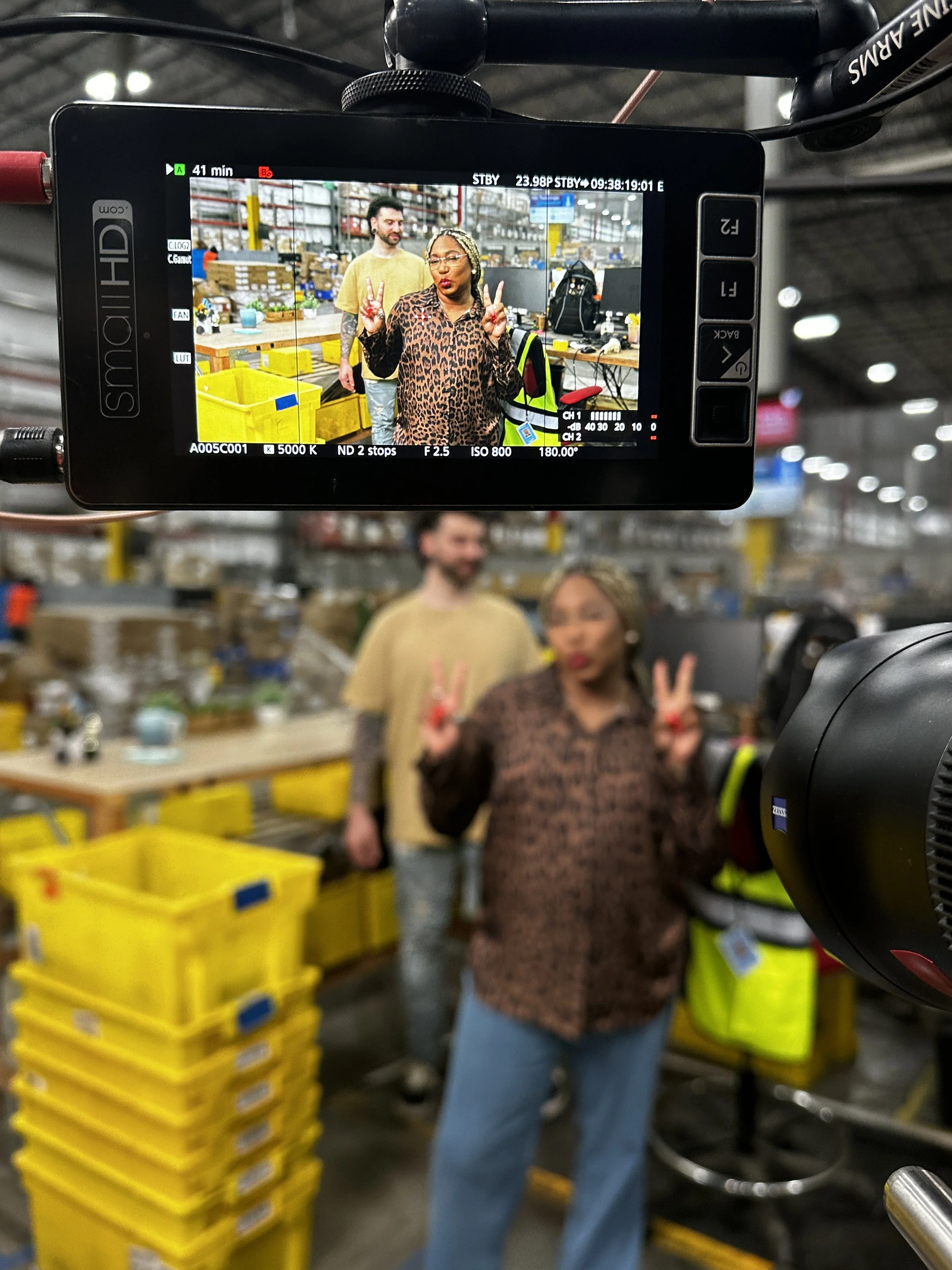 A woman wearing glasses and a leopard print jacket making peace signs in a warehouse or storage area, with two men and yellow storage bins in the background.