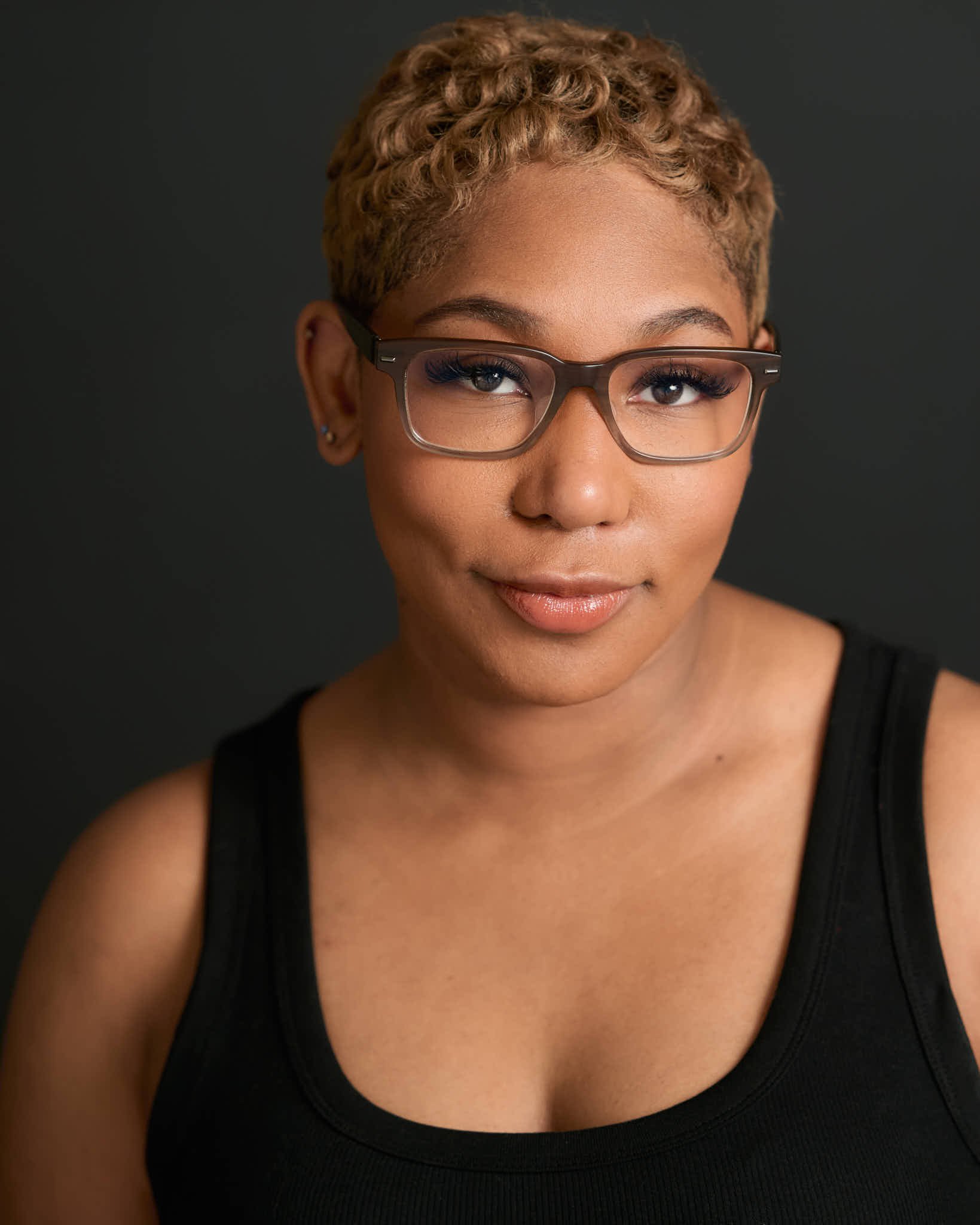 Close-up portrait of a woman with light brown, curly short hair, wearing glasses, a black sleeveless top, and natural makeup, against a dark background.