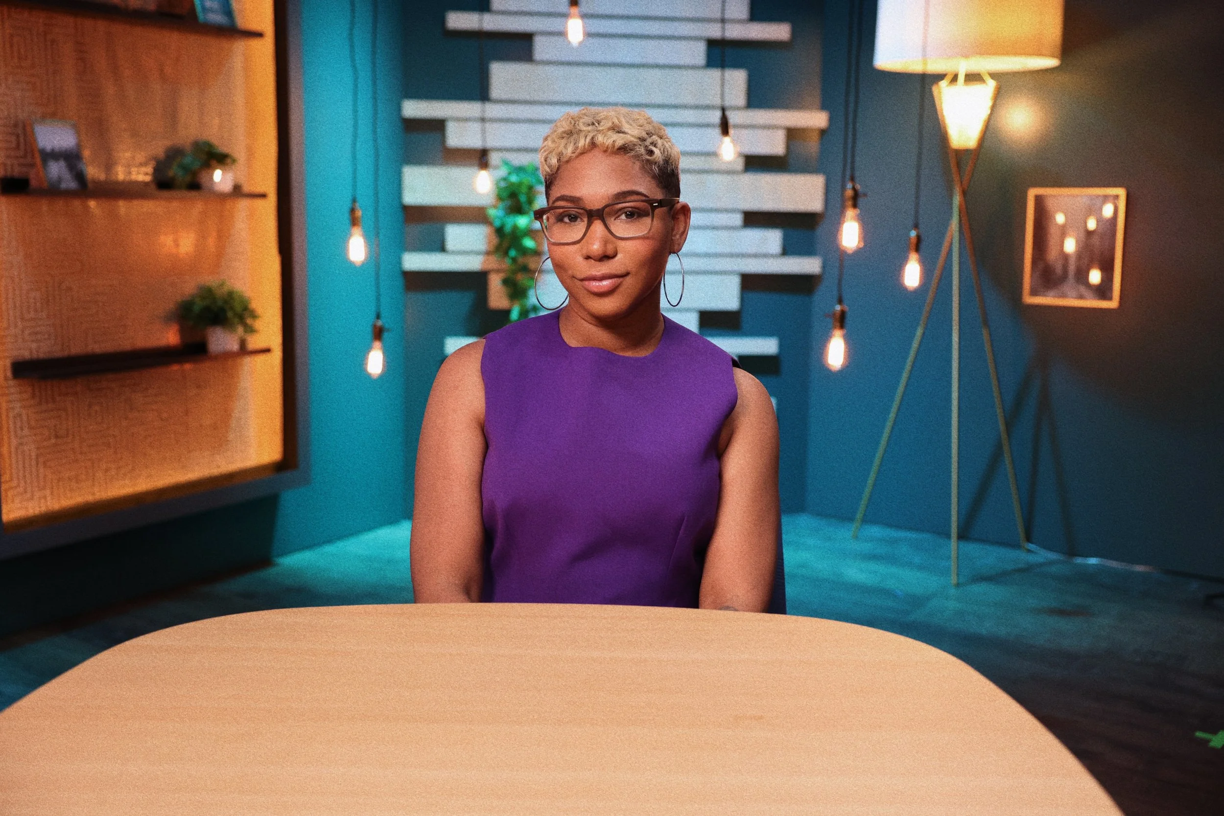 A woman with short curly blonde hair, glasses, and hoop earrings sitting at a round table in a modern interior space with blue walls, decorative lighting, and shelving with small plants.