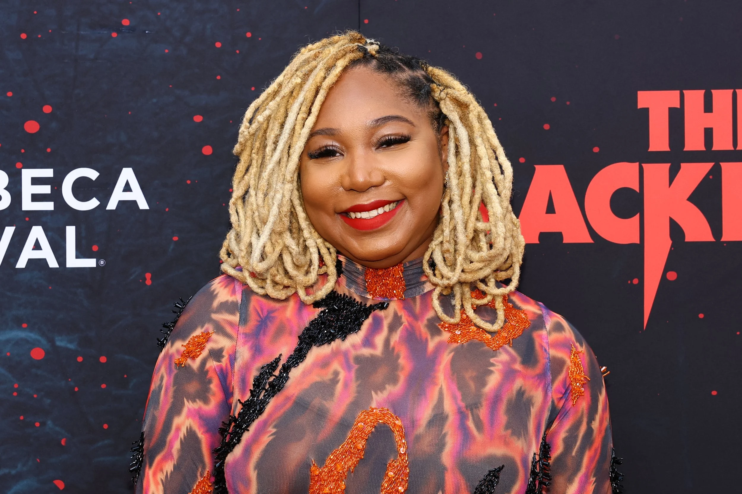 A woman with blonde dreadlocks smiling, wearing a colorful, patterned dress with orange and black embroidery, standing in front of a black background with red dots and the partial text 'THE BACK' visible.