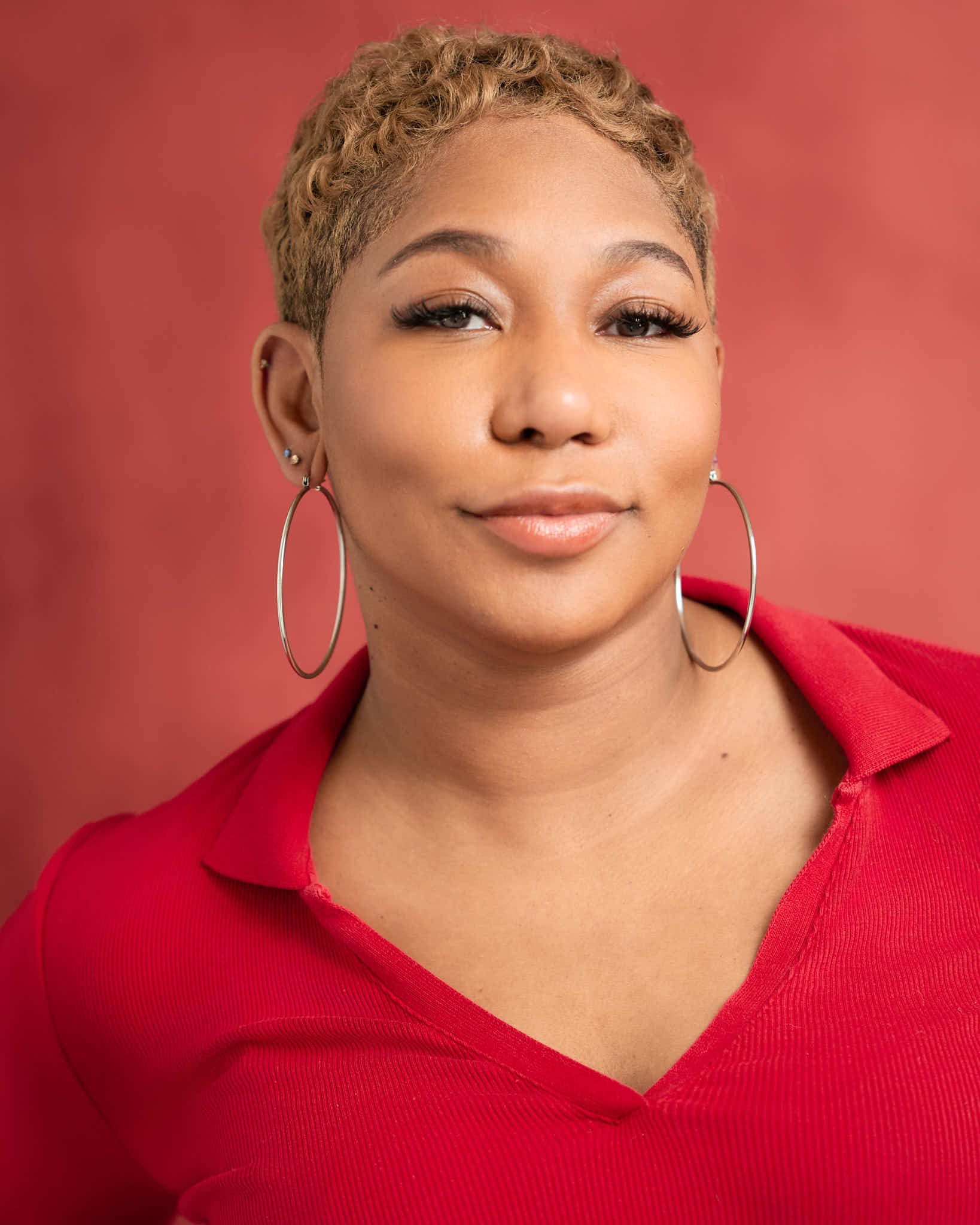 A close-up of a woman with short, curly, blond hair wearing large hoop earrings and a red collared shirt against a red background.