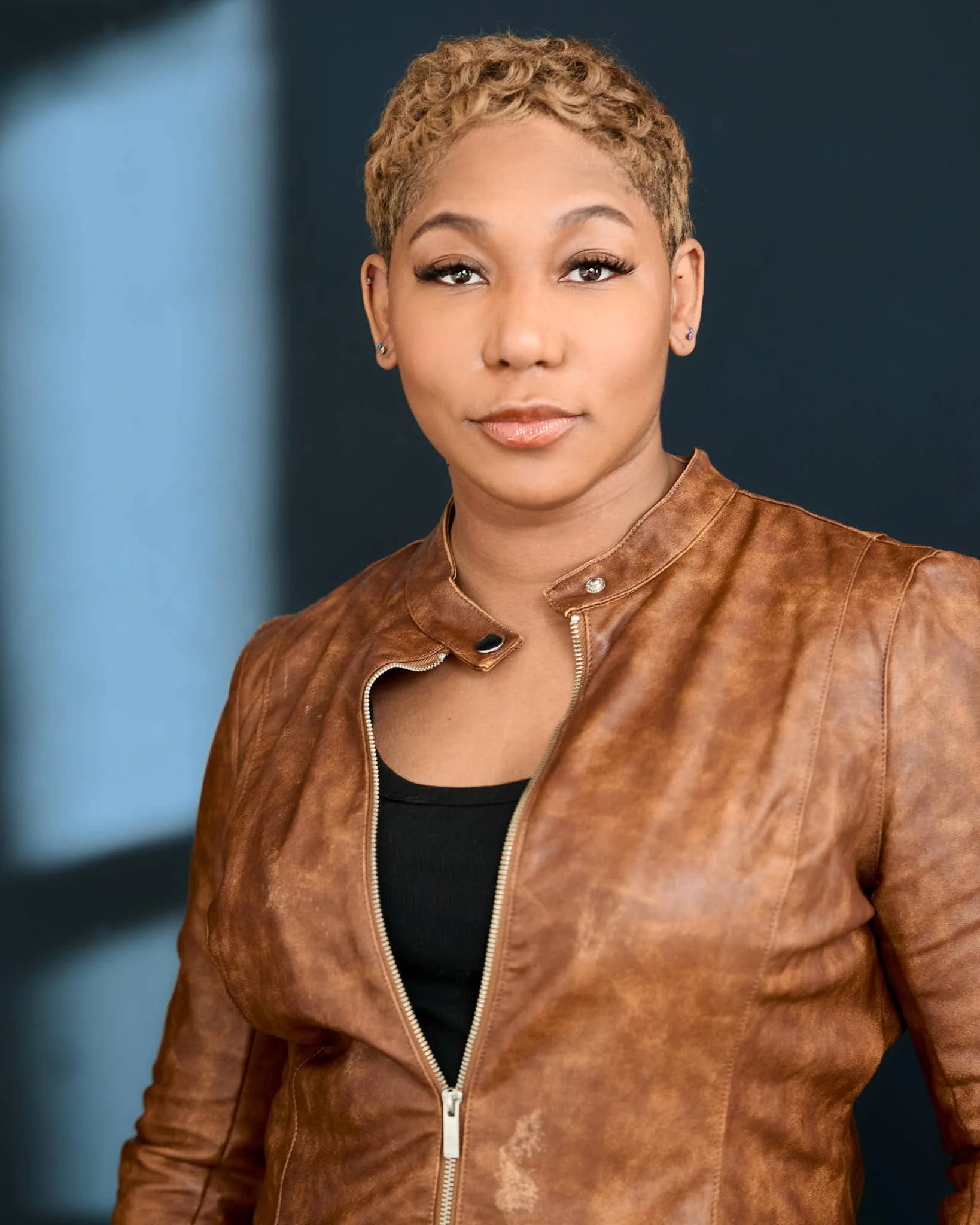 A woman with short, curly blonde hair wearing earrings and a brown leather jacket over a black top, standing against a dark blue background.