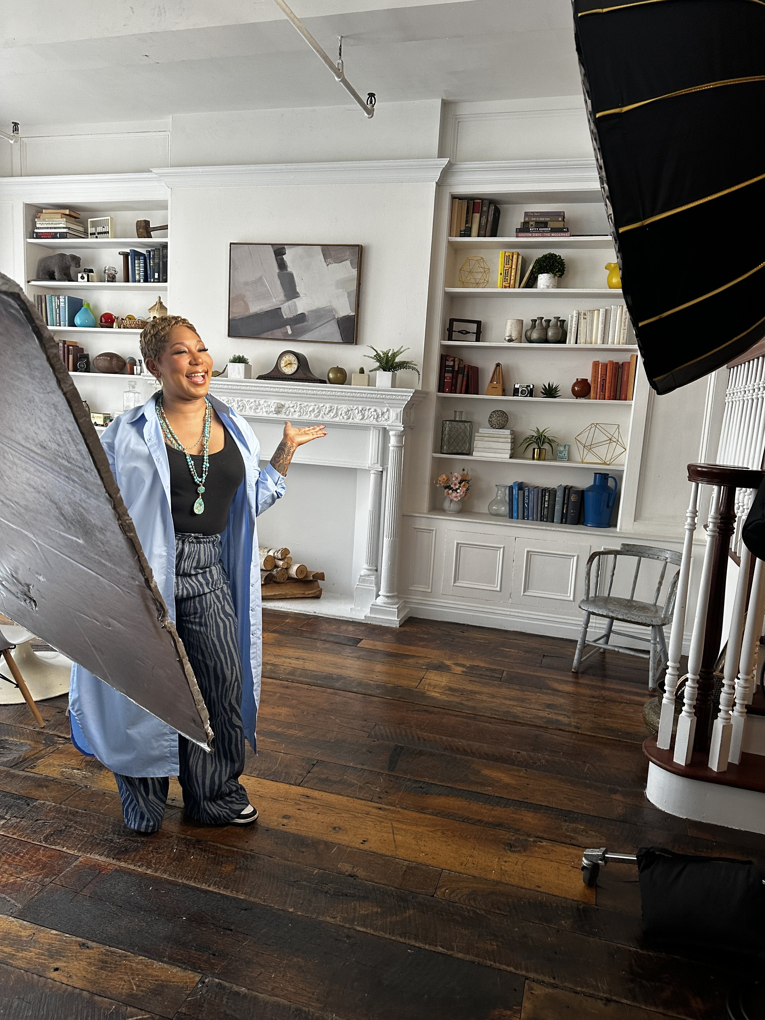 A woman smiling and gesturing indoors, with photography equipment such as a large reflector and studio lights around her, against a background of a white fireplace, bookshelves, and wooden flooring.