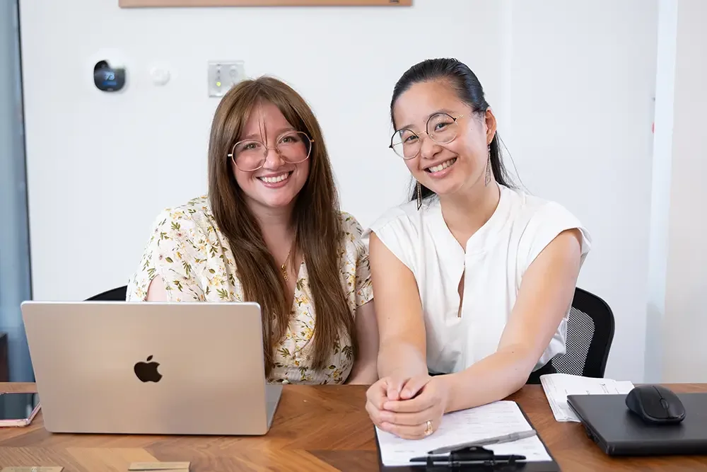 Two women sitting at a desk smiling, one with a laptop and glasses, the other with a notepad, other office supplies, and glasses.