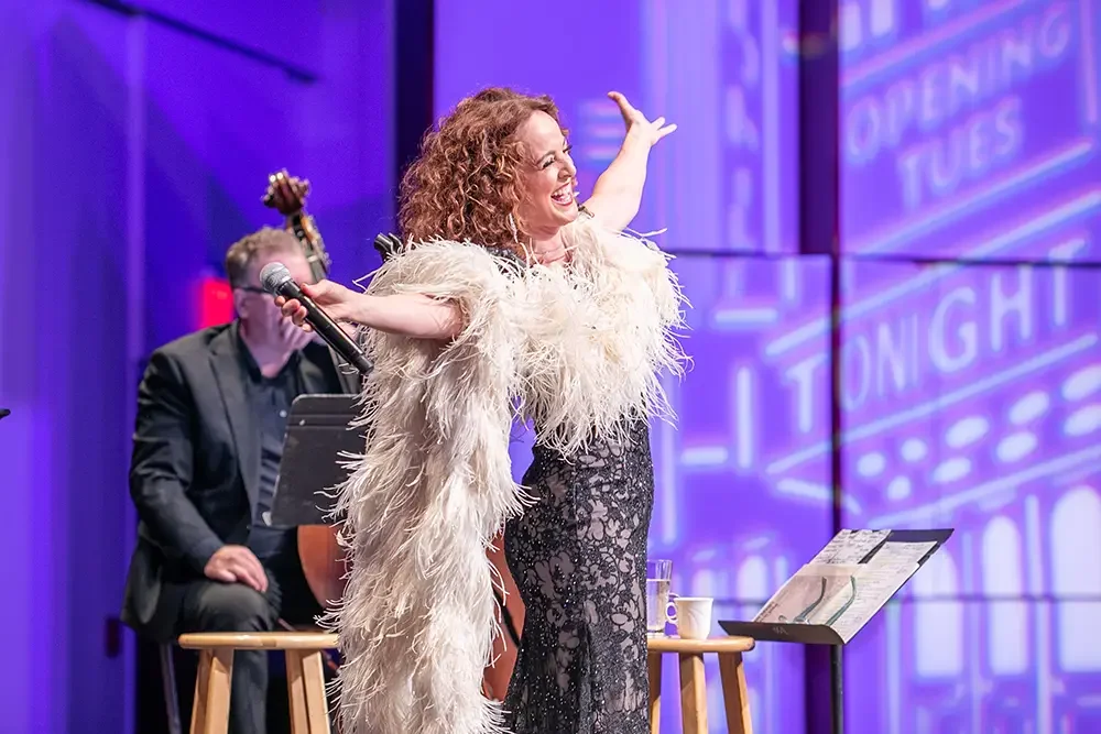 A woman with curly red hair in a glamorous black lace dress and a feathered coat is on stage, smiling and waving. A man in black suit with glasses sits behind her, playing a ukulele, on a stage with purple lighting and a background displaying text.