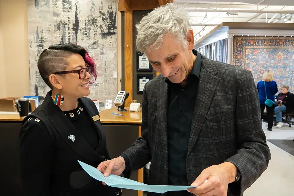 A woman with pink and black hair, glasses, and rainbow earrings is smiling at a man with gray hair, who is looking at a paper they are both holding.