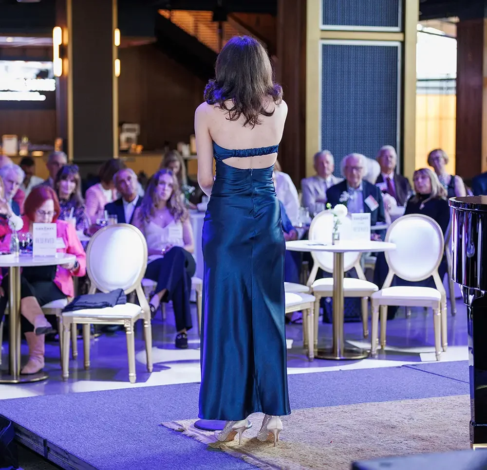 A woman in a shiny blue dress stands on a small stage in front of an audience at a formal event.