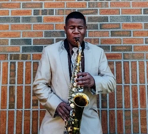 Man playing a saxophone in front of a brick wall, dressed in a light-colored suit.