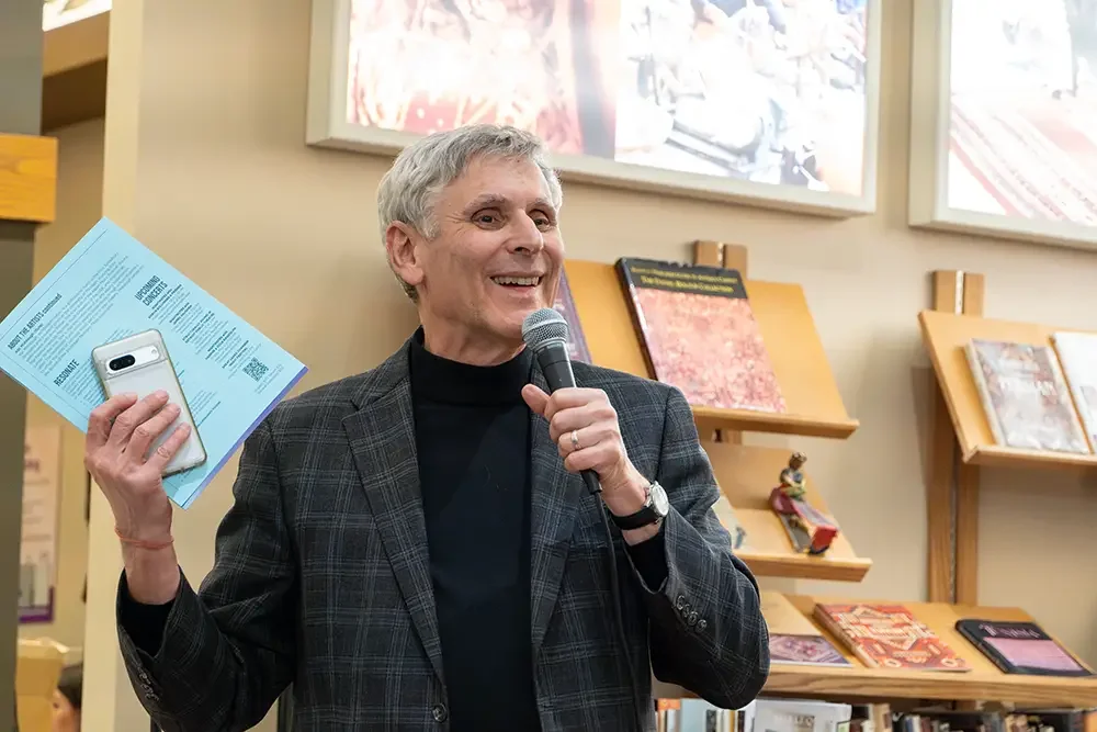 An older man with gray hair smiling while holding a microphone in one hand and a smartphone and blue papers in the other, standing in front of bookshelves and large framed artwork.