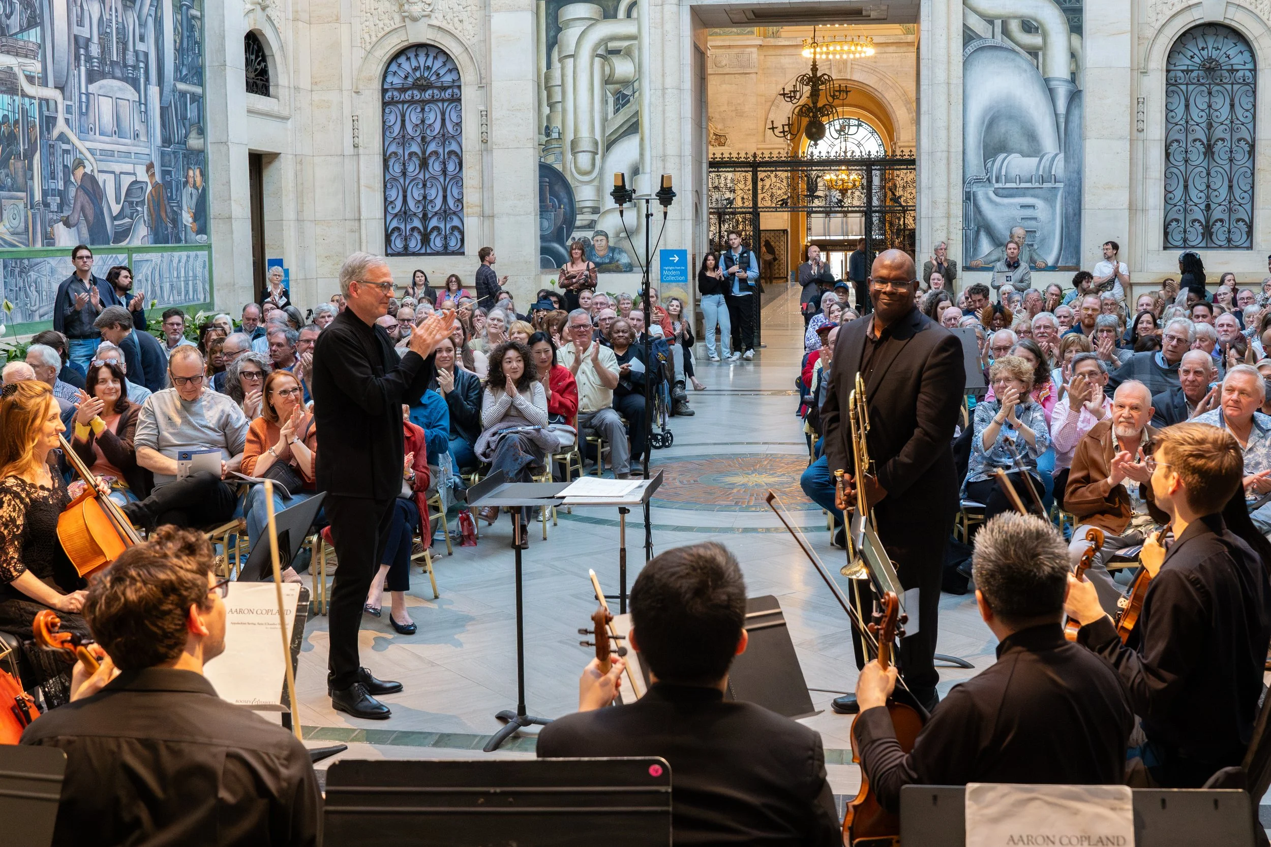 A large group of people attending a music performance inside a historic building with detailed murals and ornate architecture. The conductor stands in front of an orchestra, and a musician holding a trumpet smiles towards the audience.