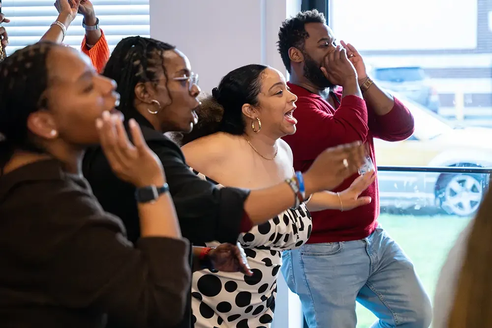 A group of diverse people watching something, some are cheering and smiling, others are surprised or thrilled, in an indoor setting with large windows.
