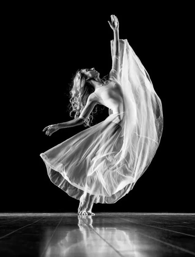 A woman wearing a flowing dress performing a dance move on stage with a black background.