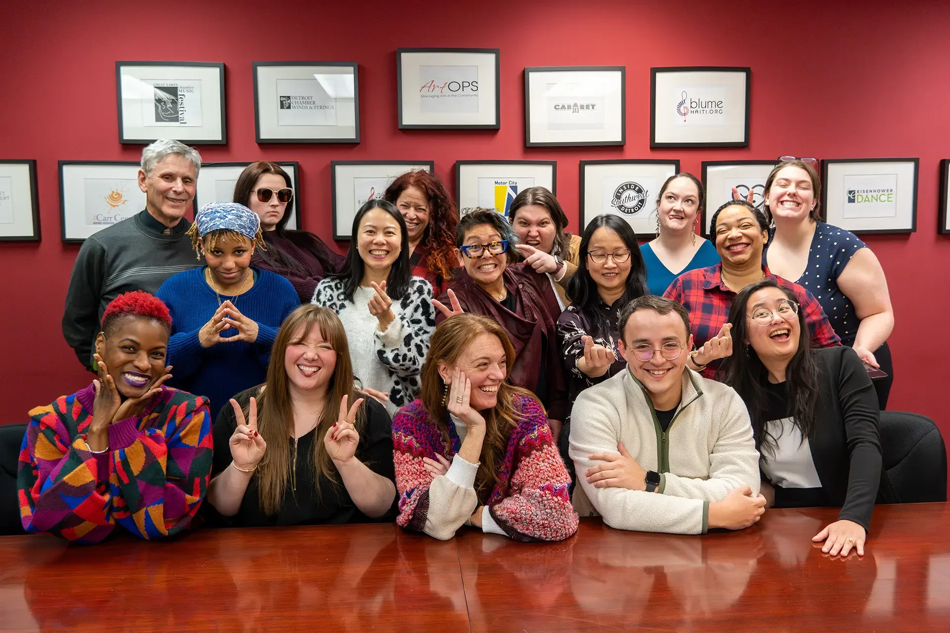Group of diverse people smiling and making peace signs in a room with framed artwork on a red wall.
