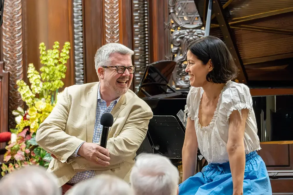 A man and woman sharing a laugh during a musical performance, with a piano and ornate wooden wall in the background.