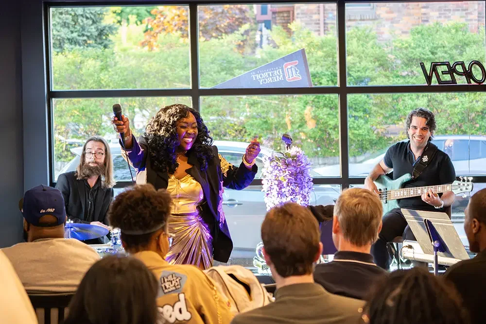 A singer performs with two musicians, one playing drums and the other playing guitar, inside a cafe or restaurant with large windows showing trees and cars outside. The audience listens attentively.