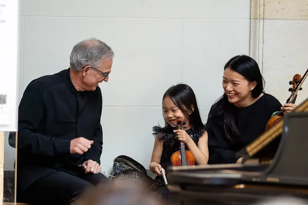 A young girl with a violin smiling with two adults, a man and a woman, also smiling, sitting near a piano.