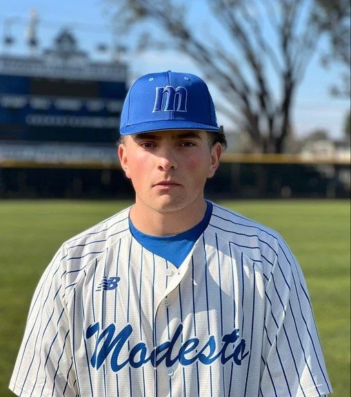 Young male baseball player in a blue cap and Michigan Modesto jersey standing on a baseball field.