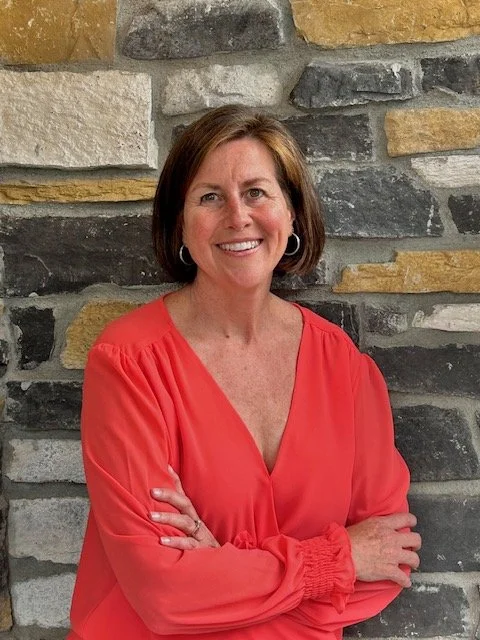 A woman with short auburn hair and hoop earrings, smiling, wearing a salmon colored blouse with long sleeves, standing in front of a multi-colored stone wall.