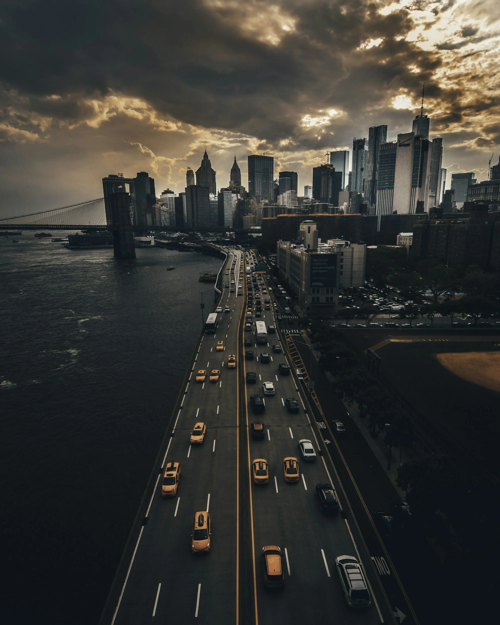 A view of the Brooklyn Bridge and the Manhattan skyline at sunset, with the East River in the foreground and busy traffic on the bridge.