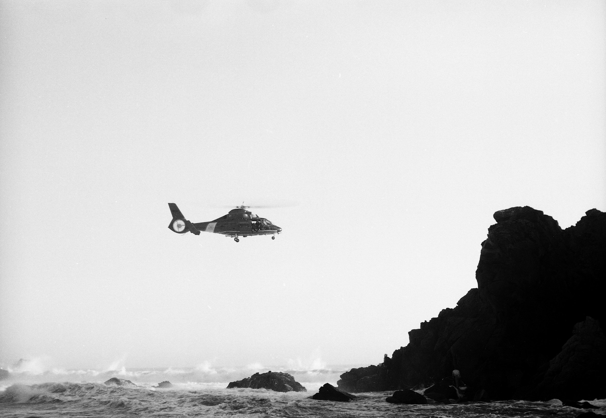 A helicopter flying over rocky coastline with ocean waves crashing against rocks.