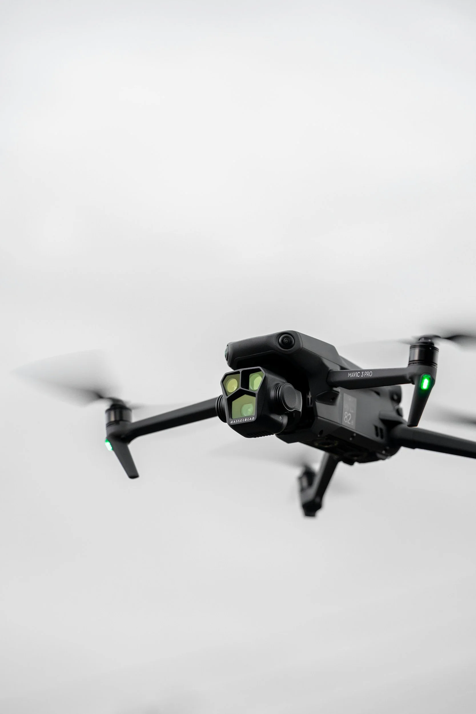 A black DJI Mavic 3 Pro drone with a camera and sensors mounted underneath, flying against a plain white ceiling.