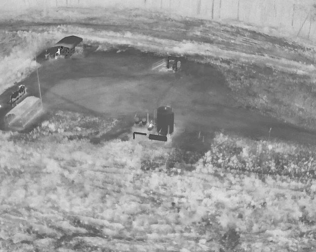 Night vision image of a field with construction equipment, including a small crane or drill, and a few vehicles, with a dirt pathway and surrounding barrier walls.