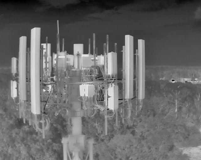 A black and white infrared photo of a telecommunications tower with numerous antennas and equipment on a grassy field.