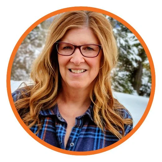 A woman with wavy blonde hair, wearing glasses and a blue plaid shirt, smiling outdoors with snow-covered trees in the background.
