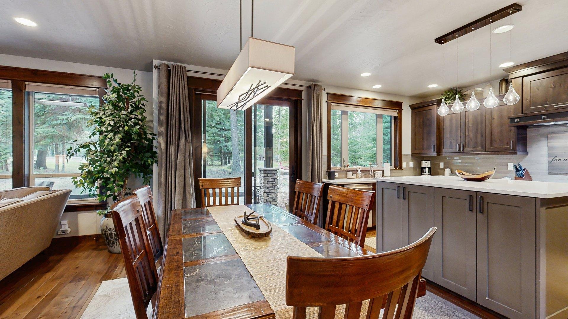Kitchen and dining area with a wooden table, eight chairs, large windows, and a sliding glass door leading to the outside. The kitchen has wooden cabinets, a white countertop, pendant lights, and a coffee maker.