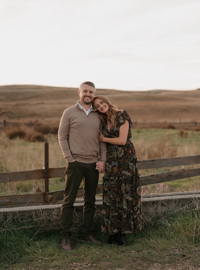 A smiling man and woman standing outdoors in a grassy field near a wooden fence, with rolling hills in the background under a cloudy sky.
