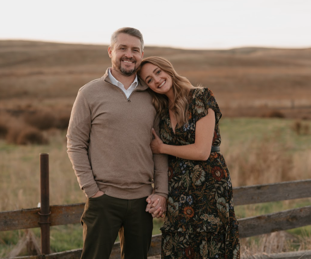 A smiling man and woman holding hands outdoors near a wooden fence with a countryside landscape in the background.