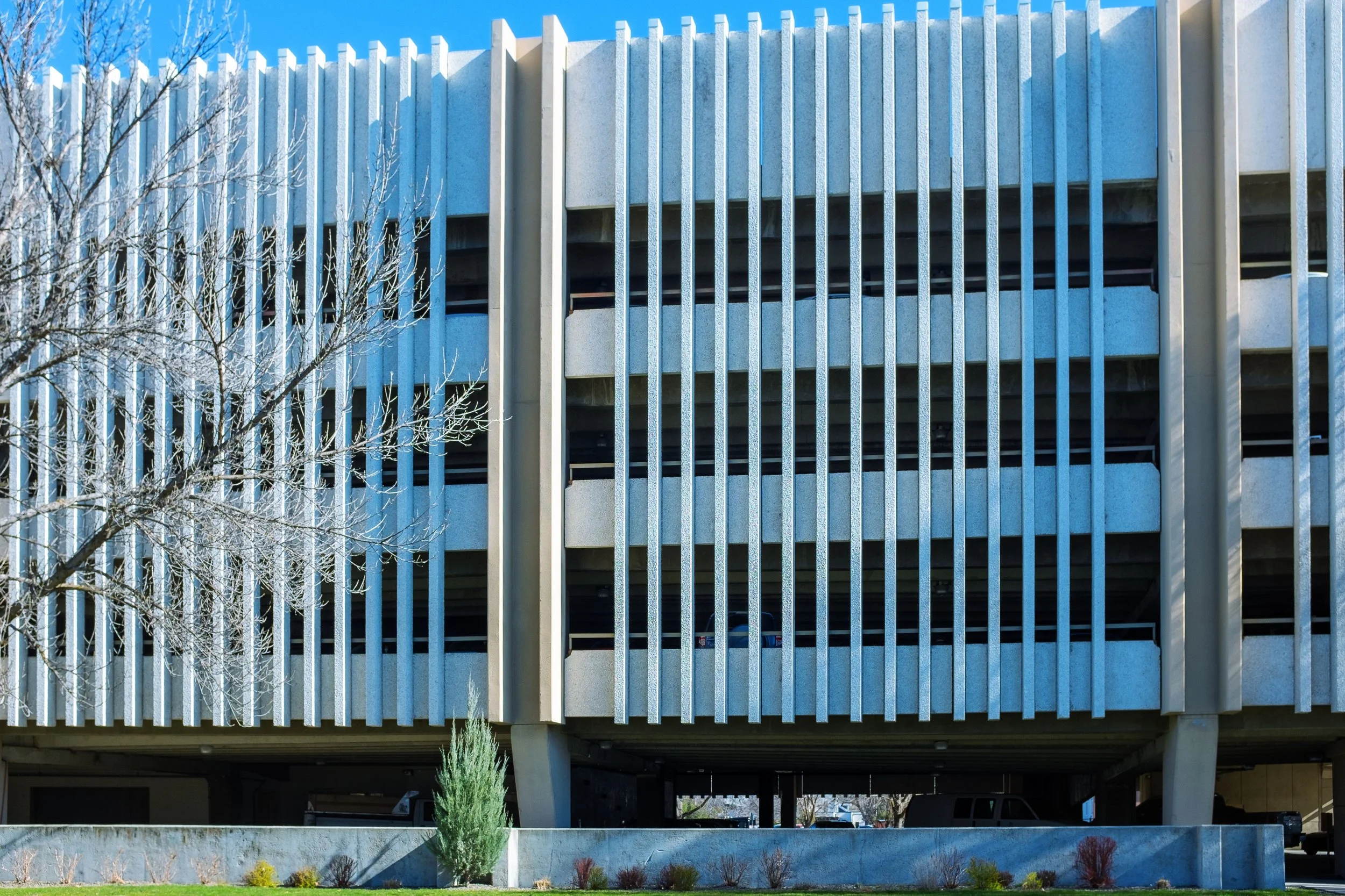 Exterior of a multi-story parking garage with vertical metal slats and some storefronts visible underneath, on a sunny day with clear blue sky and leafless trees.