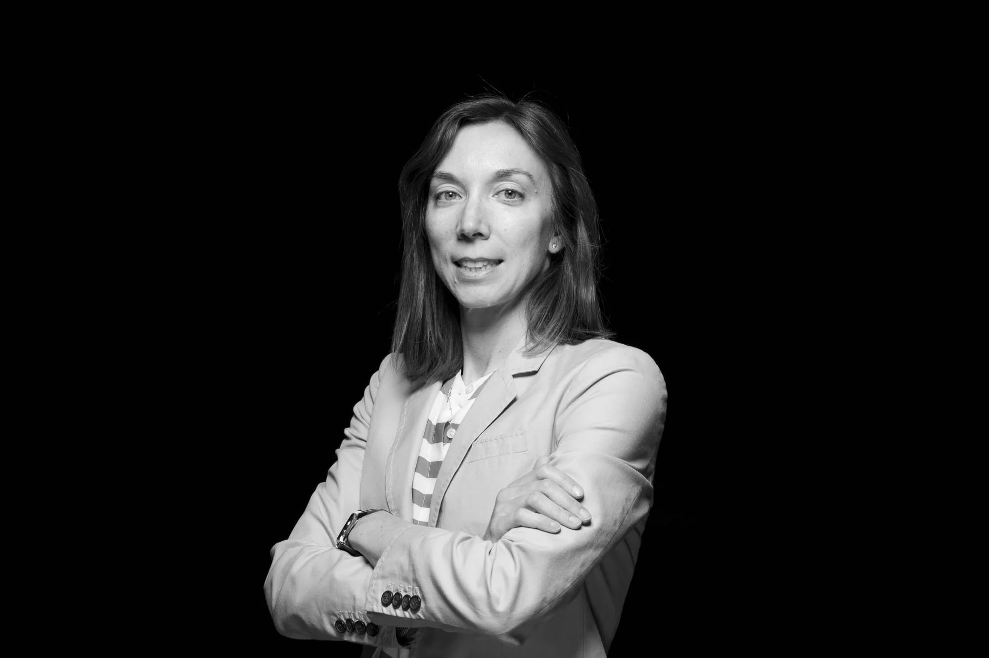 Black and white photo of a woman with shoulder-length hair, wearing a suit jacket and striped shirt, standing with arms crossed against a dark background.