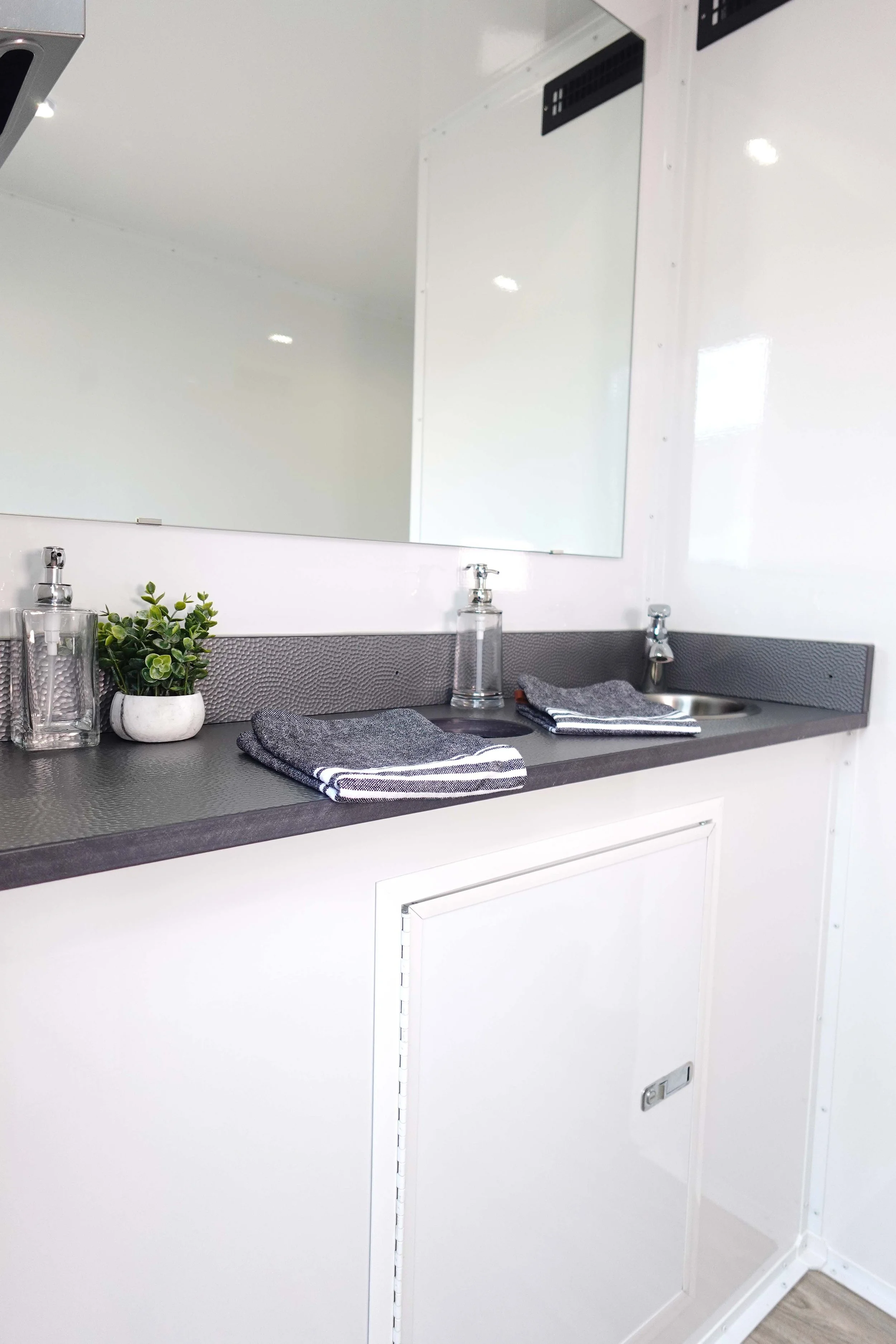 A bathroom vanity with a gray countertop, a small potted green plant, a clear soap dispenser, and a stack of gray and white towels. A large mirror is mounted above, reflecting the white walls and ceiling. There are two sinks with chrome faucets.