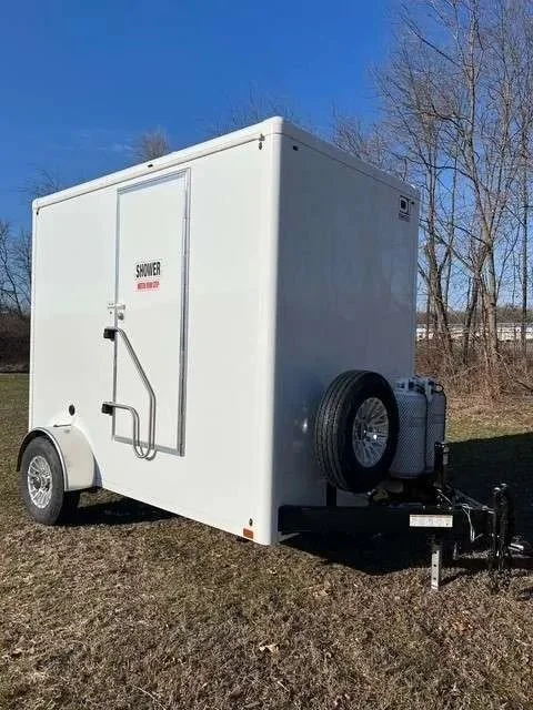 A white mobile shower trailer with a door labeled 'Shower' and a lever handle, parked on grass with a spare tire attached, under a clear blue sky.