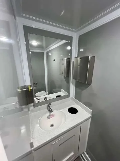 Small bathroom with a white sink, chrome faucet, and a mirror above it. There is a black cup on the sink and a wall-mounted stainless steel soap dispenser to the right of the mirror. Grey walls and a white cabinet below the sink.